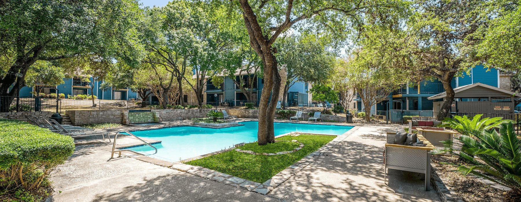 Outdoor pool area surrounded by mature trees and blue apartment buildings, with lounge chairs and patio furniture.