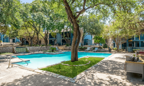 Outdoor pool area surrounded by mature trees and blue apartment buildings, with lounge chairs and patio furniture.