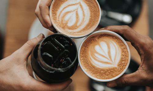 Three hands are holding cups of coffee, two with latte art and one with iced coffee, raised in a toast.