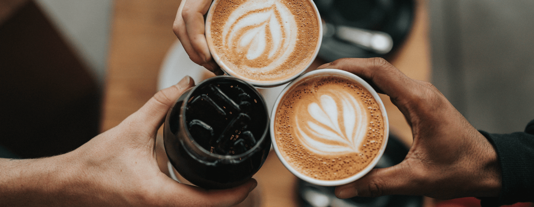 Three hands are holding cups of coffee, two with latte art and one with iced coffee, raised in a toast.