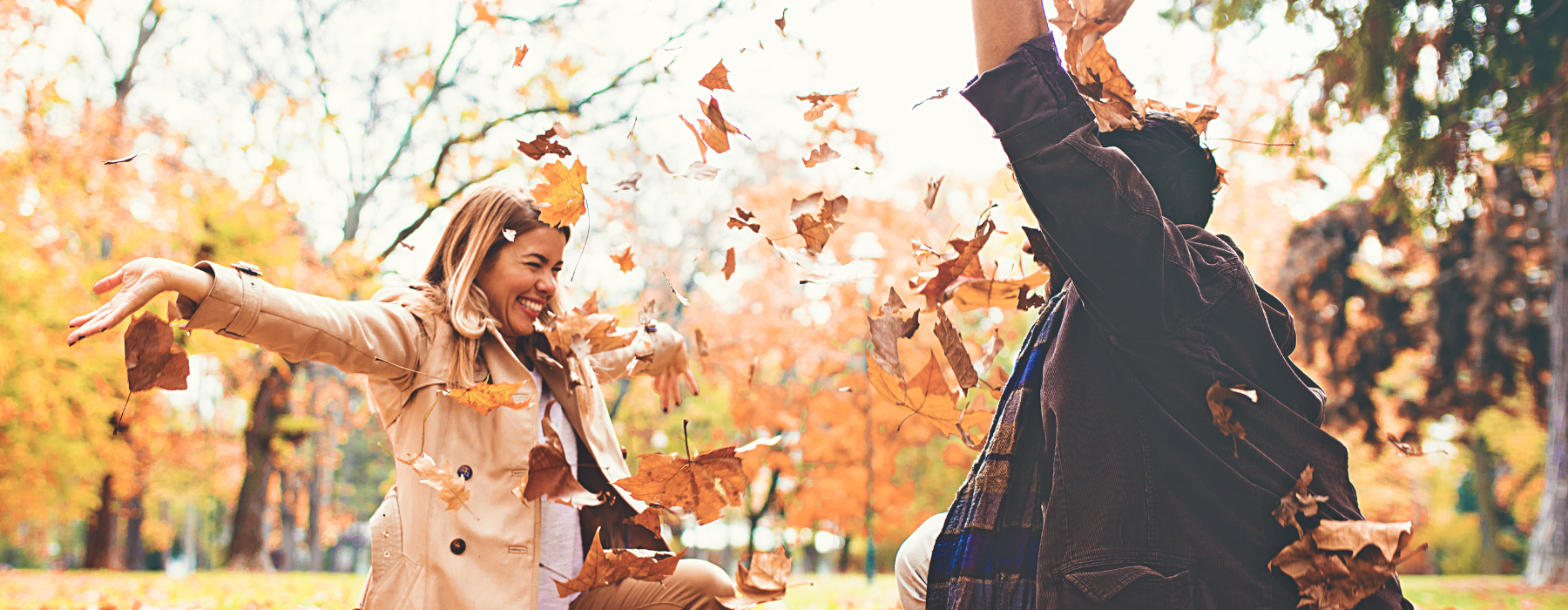  A couple enjoys a playful moment in a park filled with autumn leaves, smiling and throwing leaves in the air as the trees in the background display vibrant fall colors.