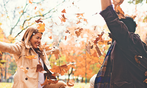  A couple enjoys a playful moment in a park filled with autumn leaves, smiling and throwing leaves in the air as the trees in the background display vibrant fall colors.