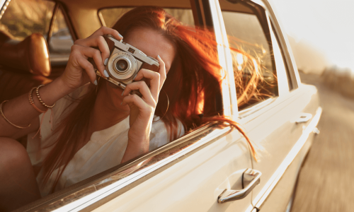 A person with long red hair is leaning out of a car window, holding a camera and taking a picture while the car is in motion.