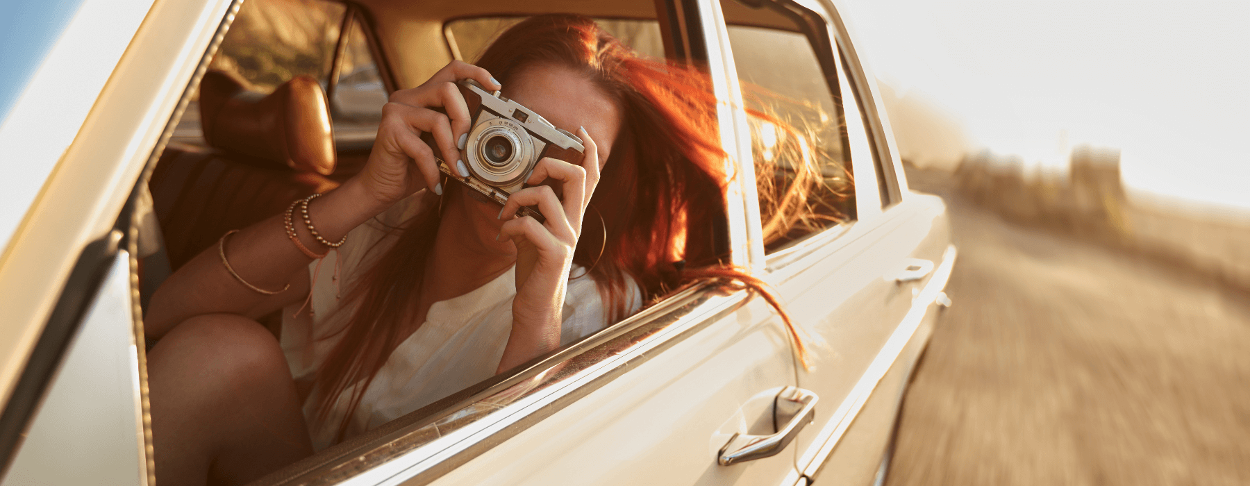 A person with long red hair is leaning out of a car window, holding a camera and taking a picture while the car is in motion.