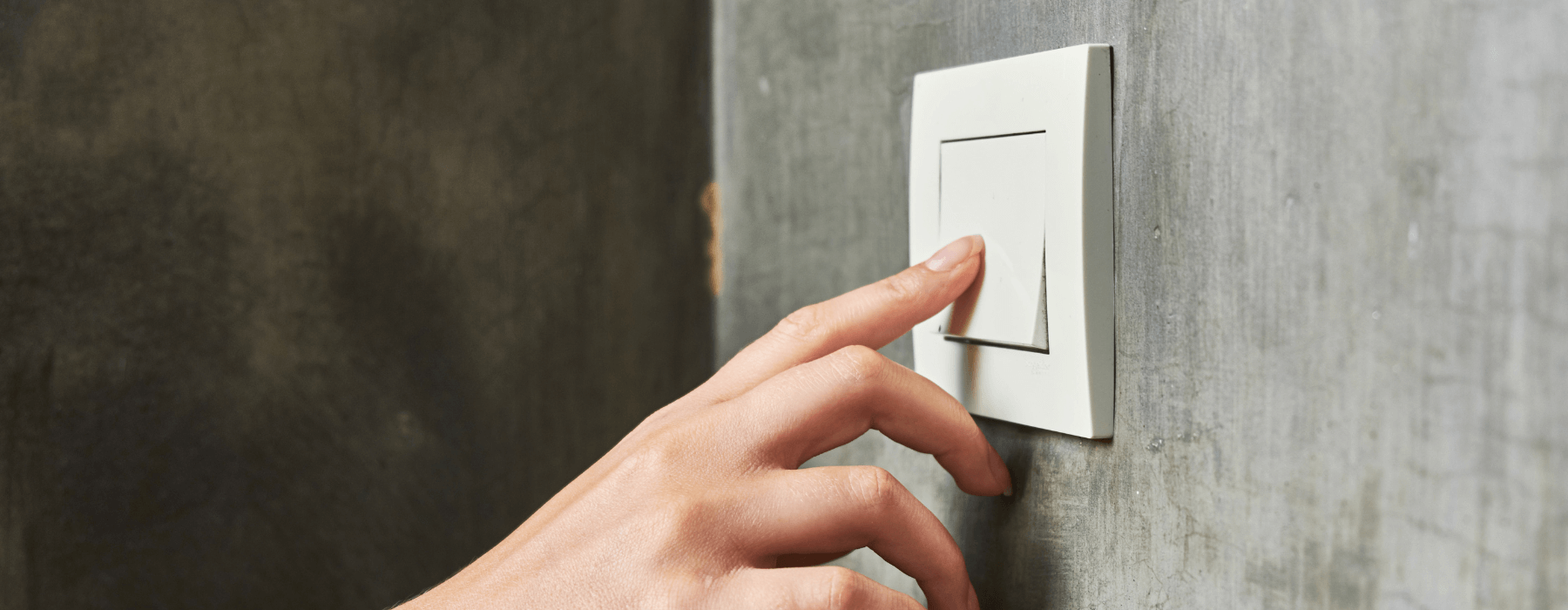 A hand is shown reaching towards and touching a light switch on a concrete wall.