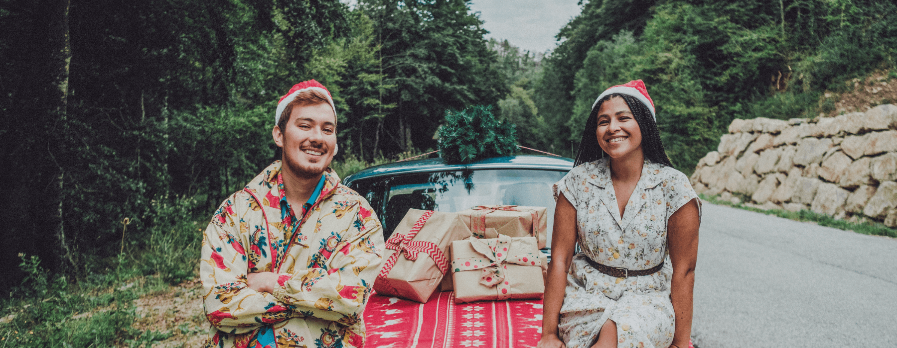 A couple wearing festive attire sitting on the hood of a car decorated with holiday presents, creating a joyful holiday scene in nature.