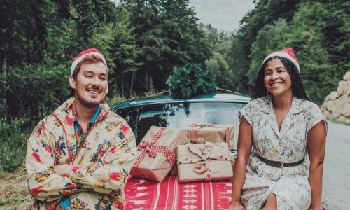 A couple wearing festive attire sitting on the hood of a car decorated with holiday presents, creating a joyful holiday scene in nature.