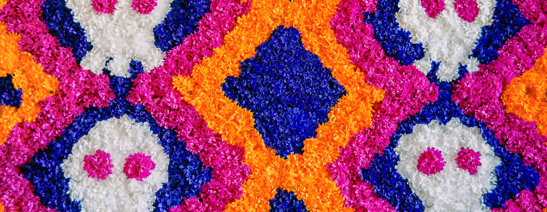 A vibrant and colorful floral arrangement with pink, orange, blue, and white flowers forming patterns, including stylized skulls, resembling Day of the Dead decorations.