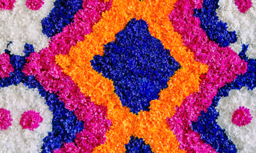 A vibrant and colorful floral arrangement with pink, orange, blue, and white flowers forming patterns, including stylized skulls, resembling Day of the Dead decorations.