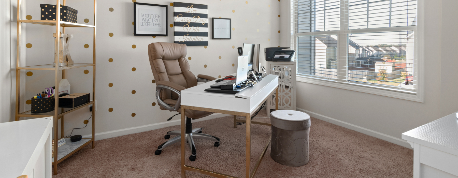 A cozy home office with a white desk, leather chair, computer setup, and gold polka dot decor. Motivational wall art and a gold shelf with office supplies complete the space.