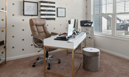 A cozy home office with a white desk, leather chair, computer setup, and gold polka dot decor. Motivational wall art and a gold shelf with office supplies complete the space.