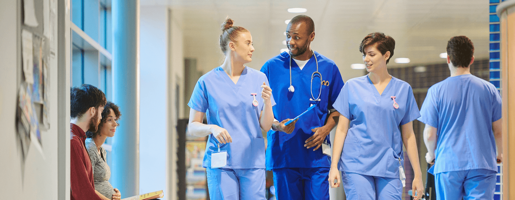 A group of healthcare professionals in scrubs conversing in a hospital corridor, with patients in the background.