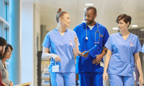 A group of healthcare professionals in scrubs conversing in a hospital corridor, with patients in the background.
