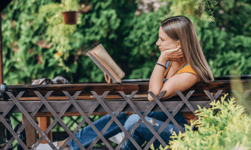 A woman sitting on an outdoor bench, surrounded by greenery and reading a book in a peaceful garden setting, enjoying a quiet moment.