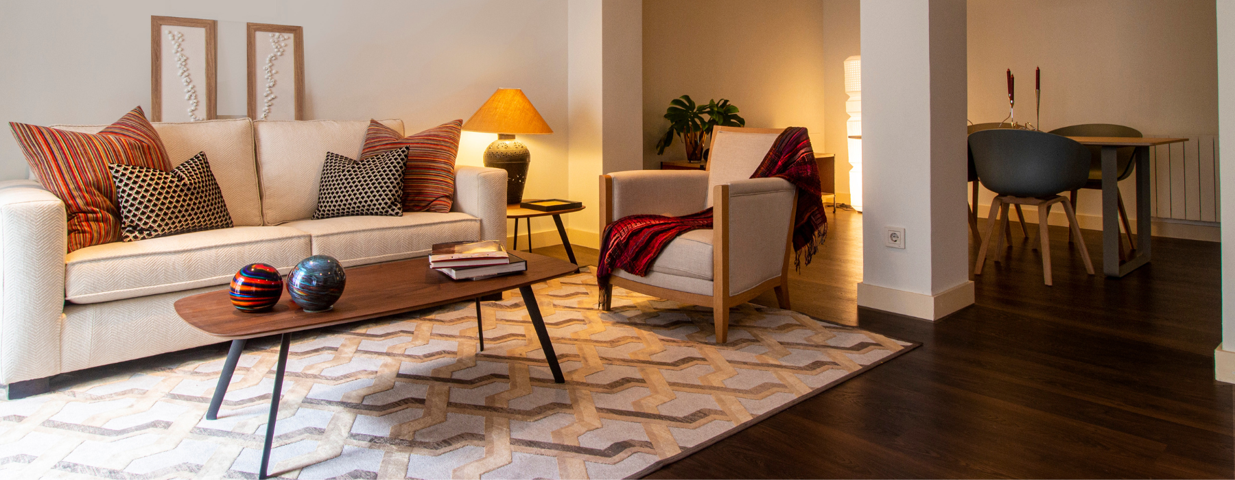 A cozy living room with a beige sofa, colorful pillows, a coffee table, and an armchair, leading to a dining area.