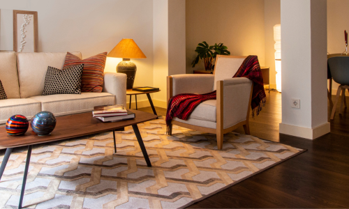 A cozy living room with a beige sofa, colorful pillows, a coffee table, and an armchair, leading to a dining area.