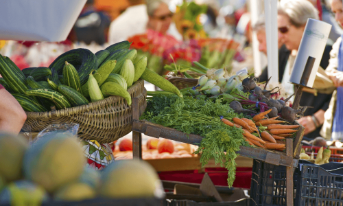 Fresh produce including cucumbers, carrots, and beets displayed at a bustling farmers market with people browsing in the background.