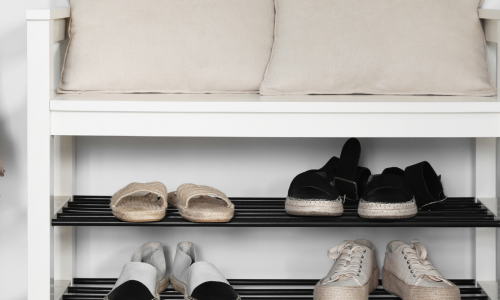 A neatly organized entryway with a cushioned bench, a tan backpack on a hook, and shelves of shoes in monochrome tones.