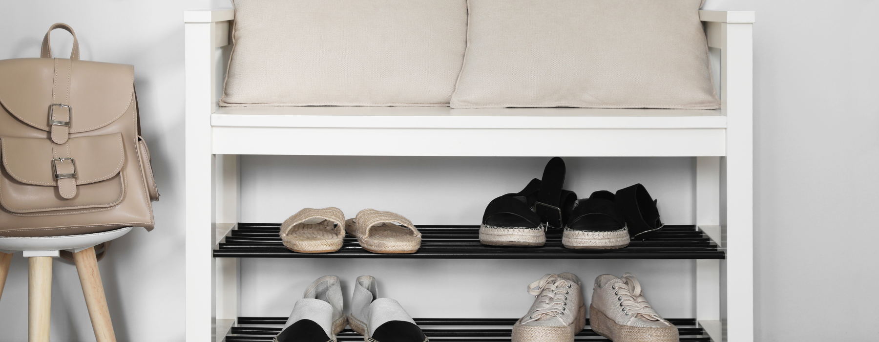 A neatly organized entryway with a cushioned bench, a tan backpack on a hook, and shelves of shoes in monochrome tones.