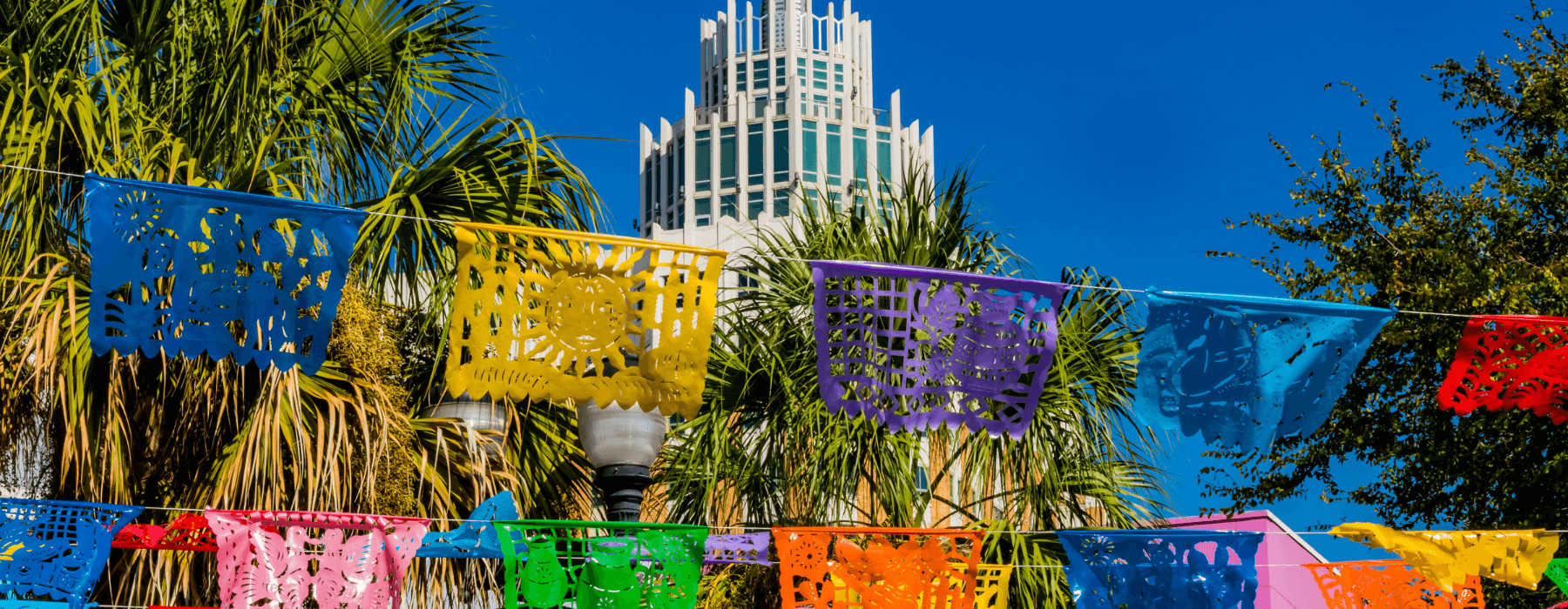 Colorful papel picado, traditional Mexican cutout banners, strung across a clear sky with palm trees and a tall, modern building in the background.