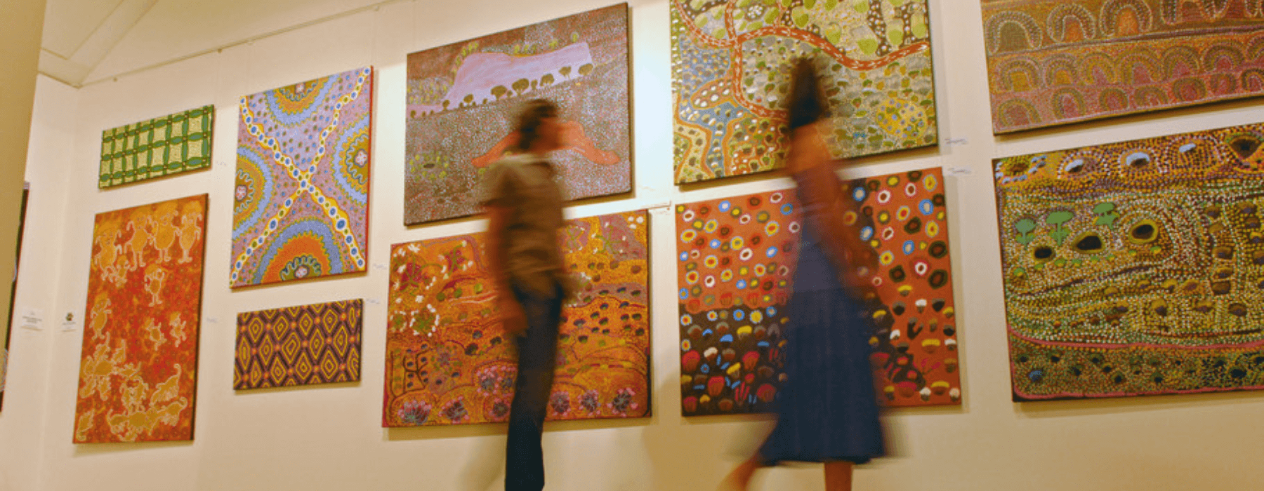 People viewing a collection of colorful and patterned art paintings on display in an art gallery.