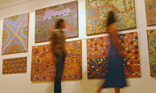 People viewing a collection of colorful and patterned art paintings on display in an art gallery.