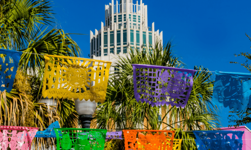 Colorful papel picado, traditional Mexican cutout banners, strung across a clear sky with palm trees and a tall, modern building in the background.