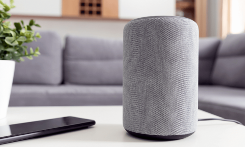  smart speaker sits on a coffee table next to a smartphone and a potted plant, set against the backdrop of a modern living room.