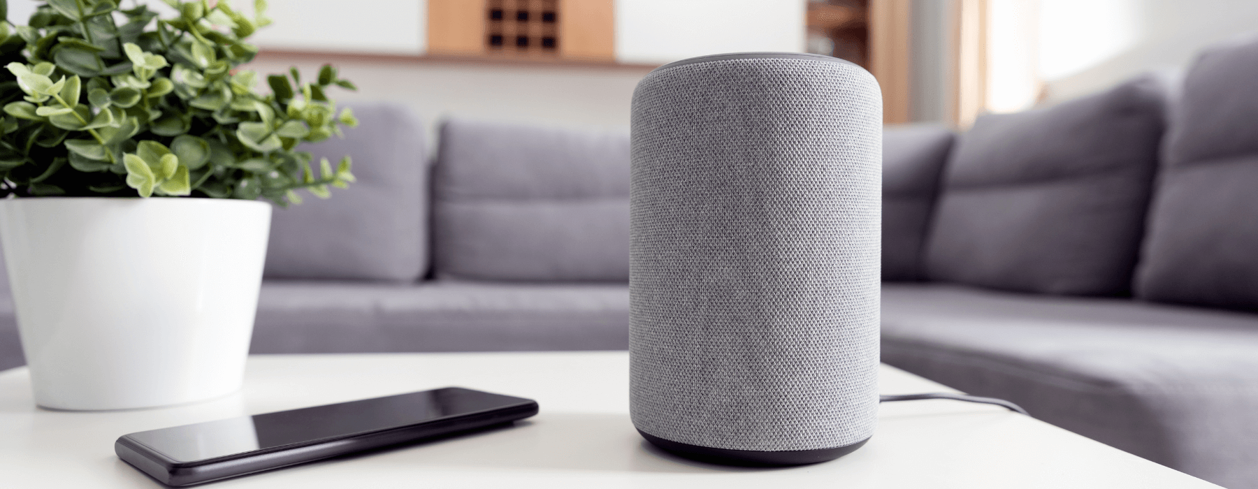  smart speaker sits on a coffee table next to a smartphone and a potted plant, set against the backdrop of a modern living room.