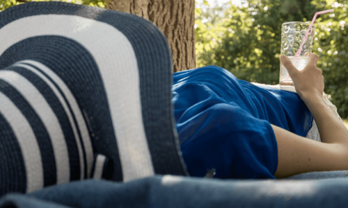 A person lounging on a blanket outdoors under the shade of a tree, wearing a wide-brimmed hat and holding a refreshing drink with a straw, enjoying a peaceful moment in nature.