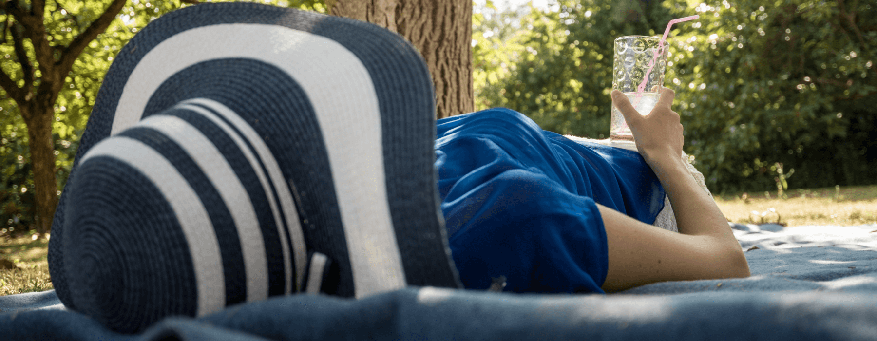 A person lounging on a blanket outdoors under the shade of a tree, wearing a wide-brimmed hat and holding a refreshing drink with a straw, enjoying a peaceful moment in nature.