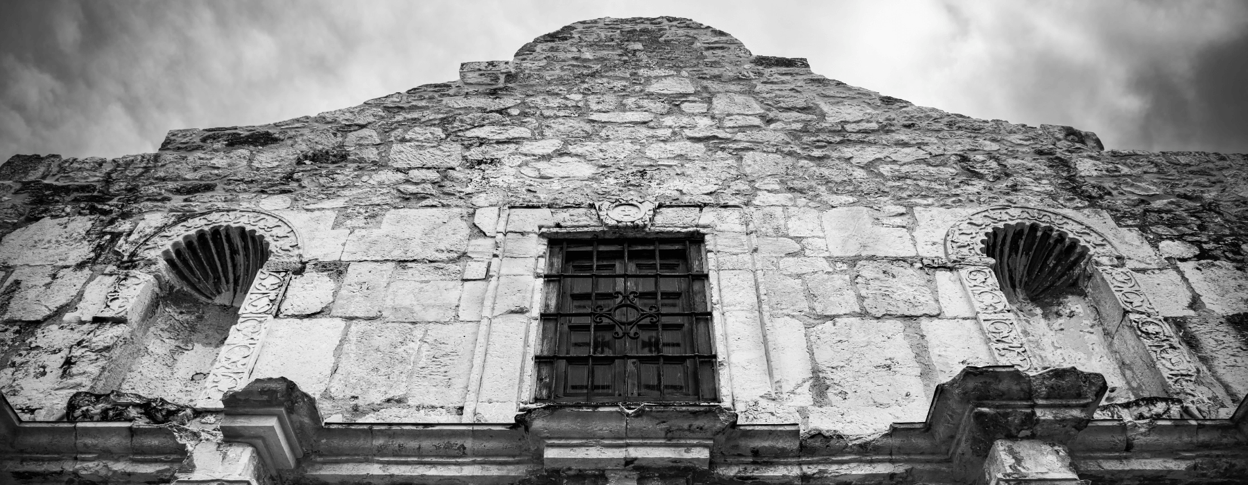 A black and white image of the upper facade of the historic Alamo Mission in San Antonio, Texas, showing its detailed stone architecture and barred window.