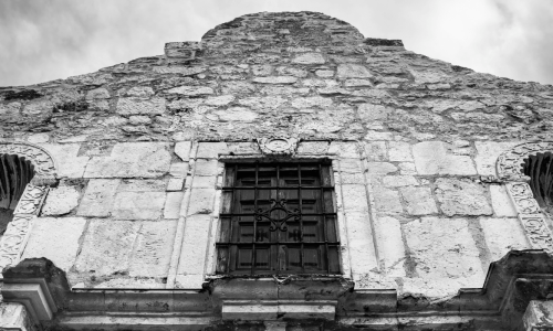 A black and white image of the upper facade of the historic Alamo Mission in San Antonio, Texas, showing its detailed stone architecture and barred window.