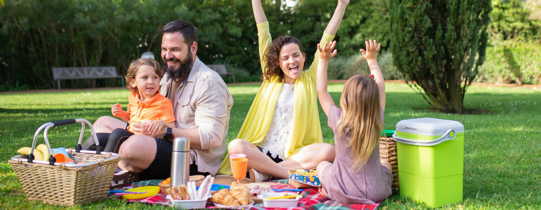 A family enjoys a picnic in a park. The father holds their son, while the mother and daughter raise their arms in excitement. A picnic basket and cooler are on the blanket.