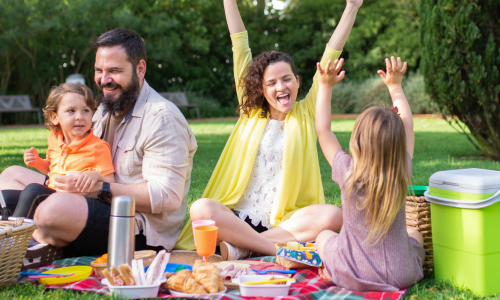 A family enjoys a picnic in a park. The father holds their son, while the mother and daughter raise their arms in excitement. A picnic basket and cooler are on the blanket.